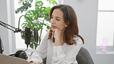 Thoughtful hispanic woman in white blouse podcasting in a modern studio with plants