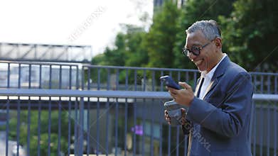 Cheerful Smiling Asian Man Walking on the Bridge Holding an Eco Cup and Using the Smartphone.