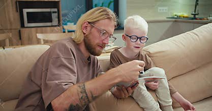 Happy little albino boy in blue glasses sits on the sofa and his dad, a blond man with long hair and a beard, explains