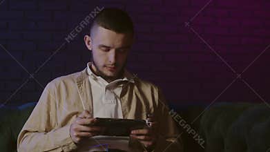 A young guy playing video games in portable console indoors. Male enjoy mobile gaming using tablet. Addicted console