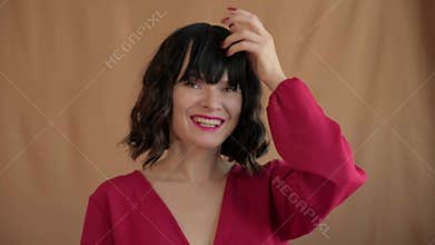 Beautiful woman smiles broadly, touching hair bangs. She strikes relaxed, spontaneous pose in red blouse. Open expression and