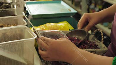 Closeup hands bagging dried hibiscus at market counter, vendor scooping petals into clear plastic bag, scale and bins visible
