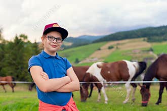 Smiling young girl in glasses stands with arms crossed in a green pasture. Horses graze behind her among rolling hills,