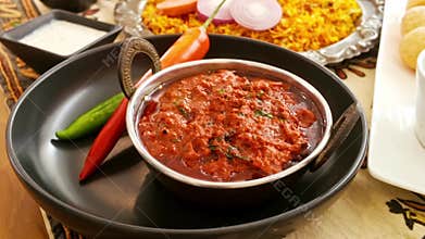 Spicy Indian curry in a metal bowl with red and green chilies, served beside saffron rice and raita on a rustic table