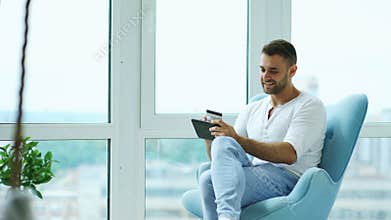 Young smiling man doing online shopping using digital tablet computer sitting at balcony in modern loft apartment