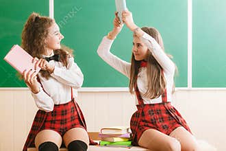 Two funny schoolgirls in school uniforms are sitting on the background of a green board on the desk with books