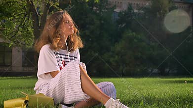 Young Caucasian brunette girl sits in a park on the grass and listens to music on headphones on a smartphone