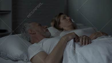 Aged man lying in bed awake, looking at woman sleeping beside him, annoyance