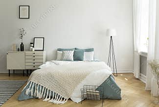 A big comfortable bed with pale sage green and white linen, pillows and blanket in a woman`s bright bedroom interior with windows