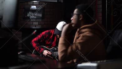 Afro-American sound producer listening to song played by Caucasian guitarist in recording studio.