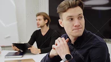 Close-up of thoughtful nervous man sitting in office with blurred positive colleague talking at background. Portrait of