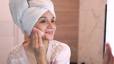 A woman with a towel on her head applies lotion to her face. a young woman with a towel on her head moisturizes her skin