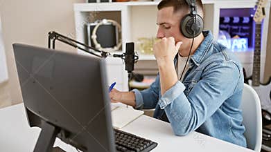 Focused young man in denim jacket podcasting with microphone and headphones at a radio studio desk