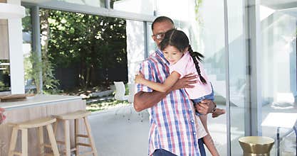 Biracial grandfather in a plaid shirt carries a young biracial granddaughter indoors