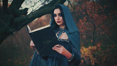 Portrait fantasy Woman witch holds book in hands looking at camera. Dark Blue dress Fairy girl vampire elf in hood cape
