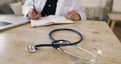 Cardiologist sits at desk, writing notes, close-up focus on stethoscope