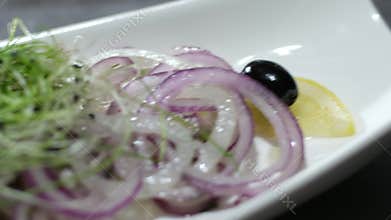 lightly salted herring on a white plate, garnished with dark olives and marinated onions with lemon