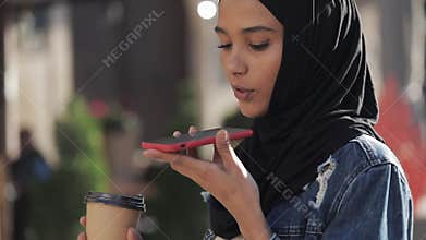 Young muslim woman sending audio voice message on cellphone at outdoor talking to mobile assistant. Girl using