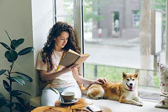 Pretty girl reading book stroking shiba inu dog relaxing on window sill in cafe
