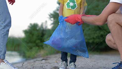 Charity cleaning, young family with little kid in rubber gloves collects refuse in garbage bag on dirty beach from