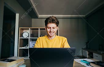 Positive guy in orange sweatshirt uses internet on laptop at home in bedroom. Cheerful student studies remotely at home, sitting