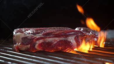 Raw beef steak falls on grill with burning fire and smoke.