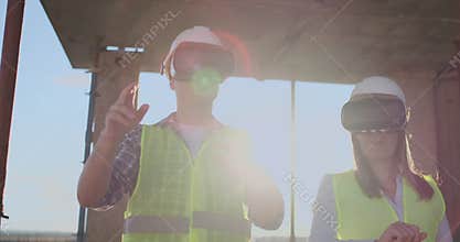 A man and a woman engineers at a construction site in VR glasses manage the construction of a building discussing a