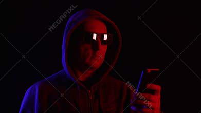 Man in sunglasses and hood using smartphone in darkness