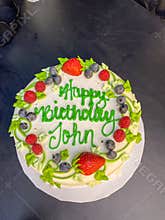 A frosted birthday cake with strawberries and blueberries that reads \"Happy Birthday John\"