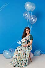 A beautiful pregnant young woman with brunette hair, sitting on the chair with balloons, on blue background.