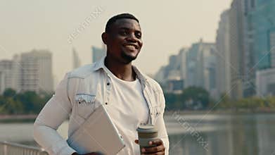 Portrait of African American man walking with to-go coffee cup on city embankment.