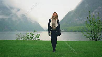 Happy woman tourist walks on green lawn near Hallstatter lake under rain.