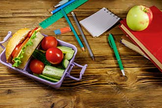 Back to school concept. School supplies, books, apple and lunch box with burgers and fresh vegetables on wooden table