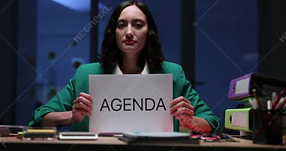 Woman holds Agenda sign sitting at desk in evening office