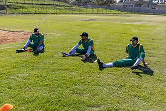 Male players sitting on grass by infield with orange cones wearing green-yellow jerseys black caps