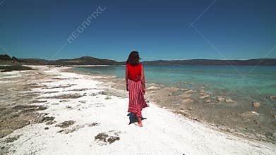 Lone woman walks along white sand beach beside turquoise water under bright coastal sun, cove, ocean, turkiye, solitude