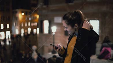 Happy young woman standing in crowd and using smartphone. Girl walking and chatting with friends in evening.
