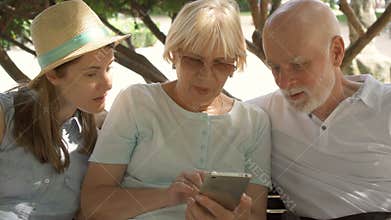 Happy family enjoying vacation. Seniors and their daughter sitting in park browsing in smartphone