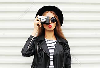 Fashion look, pretty cool young woman model with retro film camera wearing elegant black hat, leather rock jacket over white