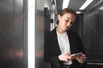 Attractive young businesswoman is looking at her smartphone srreen in the elevator while listening to music. Portrait of employee