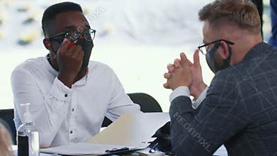 Epidemic safety at modern workplace. Happy multiethnic 20s business people talk working at office table in face masks.