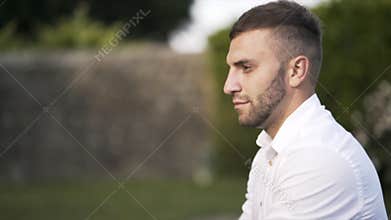 Close-up view of handsome bridegroom with brown hair and beard in white shirt looking at the camera and smiling in park