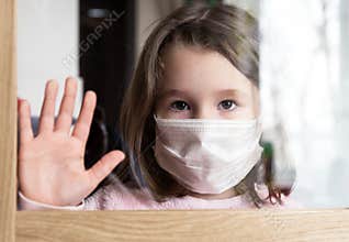 COVID-19 coronavirus concept, little girl in face mask looking through window at home or clinic. Portrait of sad kid during