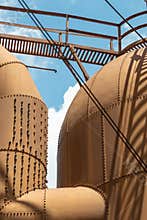 Heavily riveted metal tanks and catwalk, industrial shapes against a blue sky with clouds