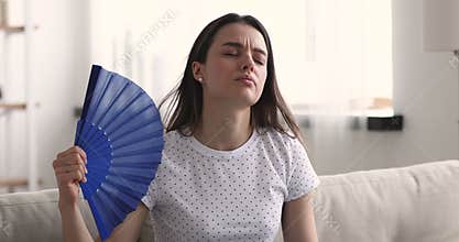 Unhappy young woman waving fan feeling uncomfortable hot at home