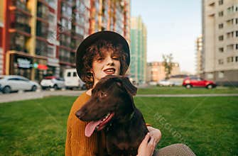 Portrait happy girl in hat sits with dog on lawn against cityscape background, hugs puppy and looks into camera with smile.Photo