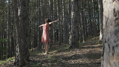 Woman in a pink summer dress walking between trees, going round with spread arms in the forest