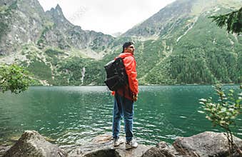 Full length portrait of a young tourist in a red raincoat and with a backpack stands against a background of a clear lake in the