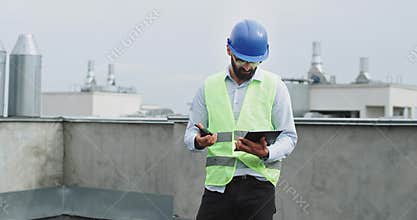 Concentrated engineer with a beard analyzing the construction plan of building on the tablet and speaking with someone