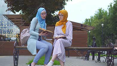 Two muslim women students in traditional scarves with textbooks in their hands communicate in the park